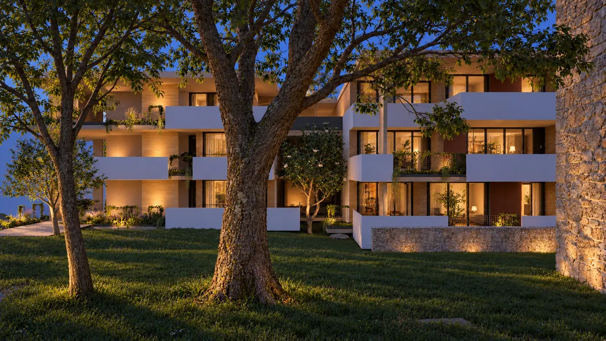 Das Haus am Abend von der Wiese aus: zwischen zwei alten Bäumen liegen drei Ebenen mit weißen Brüstungen, hinter den Fensterwänden brennt Licht; auf den Ebenen stehen bepflanzte Tröge, rechts schließt eine Wand aus Bruchstein an.