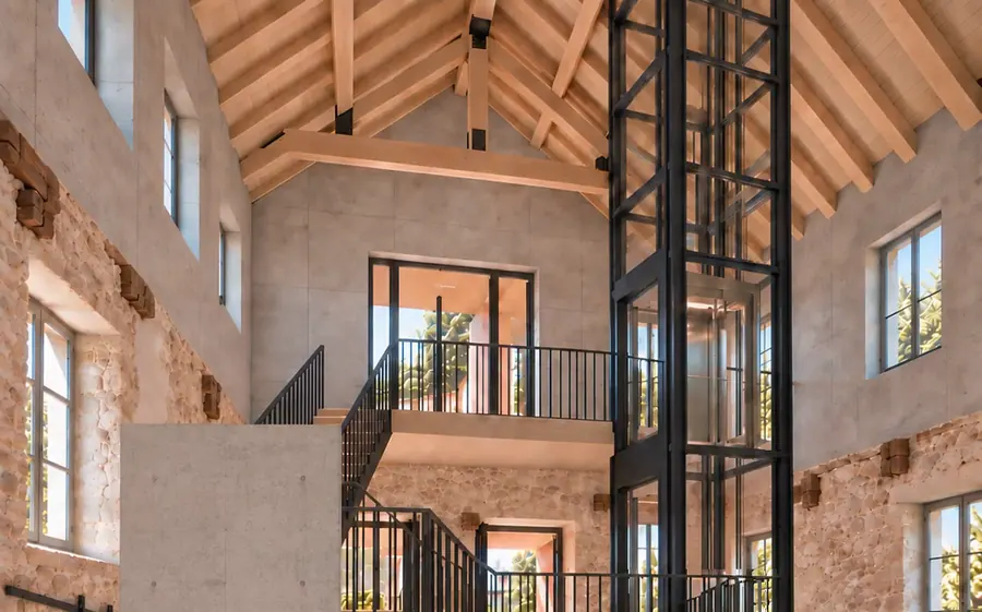 Der Aufzug im Bestand fährt zwischen den Ebenen: ein verglaster Schacht neben der Treppe, dahinter die Bruchsteinwand der alten Halle.