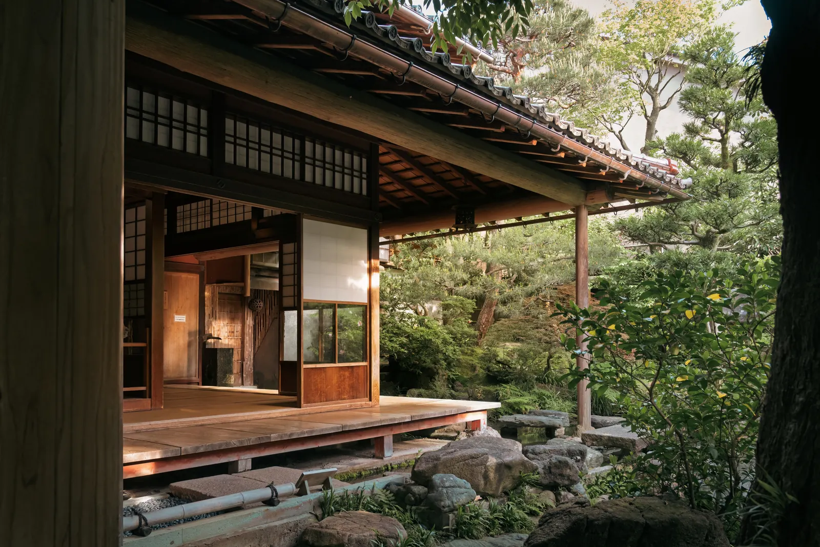 Die Veranda eines japanischen Holzhauses: unter dem tief herabgezogenen Ziegeldach ein Gang aus Holzdielen, dahinter offene Schiebetüren, davor ein Garten aus Steinen, Moos und Kiefern.