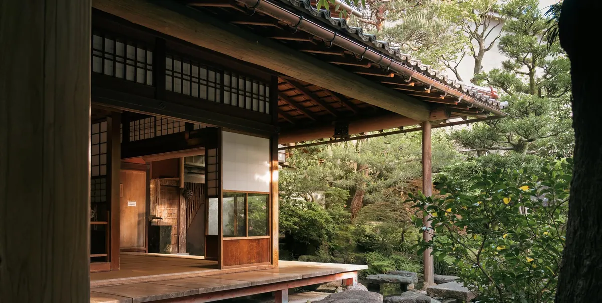 Die Veranda eines japanischen Holzhauses: unter dem tief herabgezogenen Ziegeldach ein Gang aus Holzdielen, dahinter offene Schiebetüren, davor ein Garten aus Steinen, Moos und Kiefern.